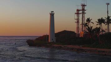 Coastal scene at sunset on Oahu island with a white lighthouse, red tower, calm ocean waves, rocky shore, palm trees, and distant mountains. video