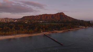 Aerial sweep of Waikiki Beach in Oahu during sunset, featuring Diamond Head, palm trees, urban structures, and a jetty extending into calm waters. video