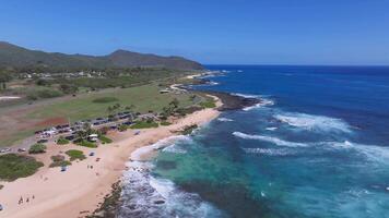 Aerial view of a Sandy beach on Oahu island, Hawaii, with turquoise waters, gentle waves, a grassy park with palm trees, and green mountains in the background. video