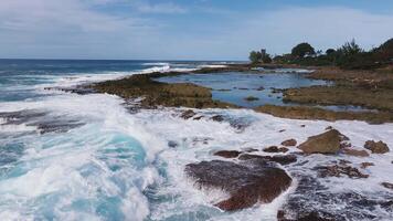Dynamic aerial view of Oahu's rocky North shore coastline with waves crashing, a small beach with visitors, and residential structures near green hills. video