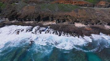 Aerial view of Oahu's coastline with turquoise waves crashing on volcanic rocks, green vegetation, reddish soil, and a road with visible vehicles. video