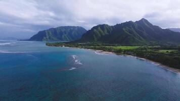 Aerial pan of Oahu, Hawaii, showing a serene coastline with turquoise waters, sandy beach, lush greenery, and mountains under sunlight breaking through clouds. video