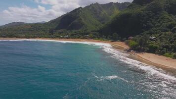 Aerial view of a Kauai beach in Hawaii with turquoise waves, golden sand, lush greenery, and forested mountains. Camera glides smoothly overhead. video