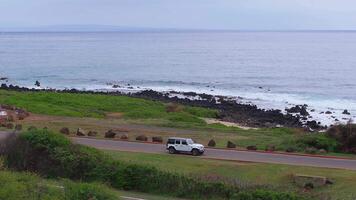A white SUV and sedan drive on a road parallel to the ocean on Oahu island, Hawaii. Grassy foreground, rocky shoreline, and calm ocean are visible. video