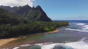 Aerial view of Kauai island's coastline, featuring a mountain, lush greenery, sandy beach, and turquoise waters with white capped waves in motion. video