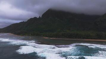 Aerial view of Napali Coast on Kauai Island, Hawaii, with lush green cliffs, mist, crashing turquoise waves, golden sand, and an overcast sky. video