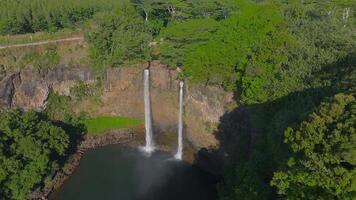 Twin waterfalls cascade over a steep cliff into a pool surrounded by lush greenery. The water flows continuously, creating mist at the base. video