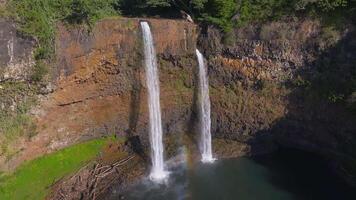 Twin waterfalls cascade from a rocky cliff into a serene pool, surrounded by lush greenery. Water flows continuously, creating mist at the base. video