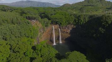 Aerial view of Wailua Falls on Kauai island, Hawaii, with twin waterfalls, lush green valley, tropical vegetation, and distant mountains in motion. video