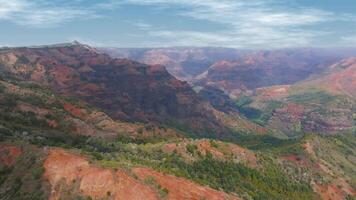 Aerial view of Waimea Canyon on Kauai island, showcasing red and green hues, steep cliffs, lush vegetation, and a clear blue sky with scattered clouds. video
