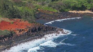 Aerial view of Kauai's coastline in Hawaii, featuring ocean waves crashing against dark volcanic rocks, red soil, and lush greenery. video