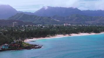Aerial view of a beach on Oahu Island, Hawaii, with turquoise waters, white sand, lush greenery, a small town, and mountains partially in clouds. video