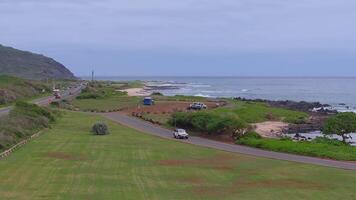 A white SUV and sedan drive on a road parallel to the ocean on Oahu island, Hawaii. Grassy foreground, rocky shoreline, and calm ocean are visible. video