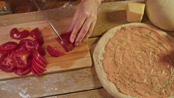 Top view of housewife carefully slicing ripe tomato into thin slices, next to pizza base made from dough seasoned with tomato paste and garlic sauce. video