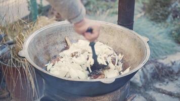 Chef mixes meat and vegetables in large cauldron with slotted spoon. Roast beef and onion in boiling oil. Tourists camp in nature. Cooking camping food in pan. video