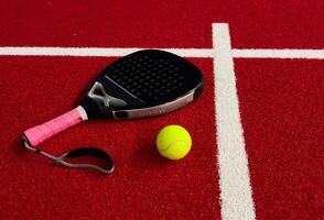 A padel or paddle tennis racket and a bright yellow tennis ball lie on a red court. The net photo