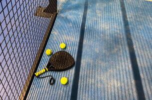 A padel or paddle tennis racket and a bright yellow tennis ball lie on a blue court. The net and a blurred figure of a player are visible in the background. photo