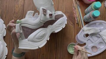 Flat lay of girl's hands paint sneakers with brush on wooden table. Designer's workshop. Palette with green acrylic and other tools on desktop. Hobbies, creativity and handmade work at home. video