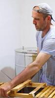Bearded man in cap pulls a frame from a big metal box. Apiarist applies electric knife to open cells on the honey frame to prepare it for extraction. Vertical video