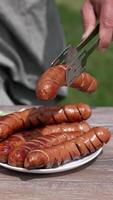 Picnic food on blur nature background. Plate with roasted sausages and fresh vegetables on table. Man adds grilled sausage using tongs to a plate. Close-up. Vertical video