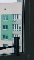 Man cleans the window and squeezes water with a brush. Male worker washing glass inside the flat. Block of flats background through clean window. Householding chores. Vertical video