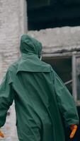 Perplexed survivor in abandoned place. Human in safety uniform and gas mask standing alone and looking to the sides on the background of destroyed buildings. Vertical video