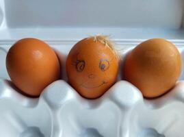 There are three brown eggs in a white package, one with a funny face and a chicken feather stuck on it. photo