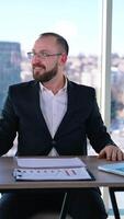 atônito homem de negocios às ambiente de trabalho. sorridente escritório trabalhador sentado às a mesa com uma computador portátil em janela fundo com cidade visualizar. vertical video