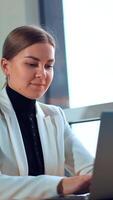 Calm Caucasian lady sitting at the desk working on laptop. Business woman typing on computer low angle view. Window at backdrop. Vertical video