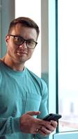 Approaching to a broad-shouldered handsome man in glasses leaning on the window frame. Portrait of a smiling man looking into camera. Close up. Vertical video