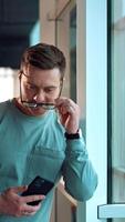 Confident good-looking man leaning on the window frame indoors. Mid-aged smiling Caucasian man puts on glasses and looks at camera. Close up. Vertical video