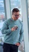 Handsome mid-aged man in glasses stands at the panoramic window with nice view. Office employee having a break from work enjoying coffee. Vertical video