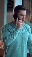 Fit mid-aged man in casual clothes stands at the window. Man looks into window and laughs speaking on the phone. Blurred backdrop. Vertical video