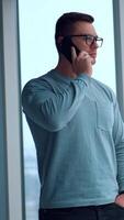 Caucasian fit man standing at the panoramic window with hand in the pocket. Businessman having phone conversation looking at the window. Vertical video