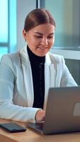 Young woman in white jacket sitting in front of her laptop. Smiling lady types something on computer and looks thoughtfully into window. Vertical video