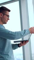 Serious confident businessman standing indoors neat the big window. Man opens his laptop typing something on the keyboard. Side view. Vertical video