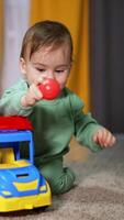 Happy adorable child with bright toy lorry on the floor in front of him. Baby boy takes a red ball out of the car basket and throws it cheerfully. Vertical video