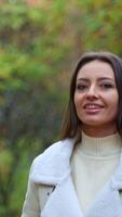 Smiling brunette in light warm jacket walking in the nature. Girl taking a stroll in the park in autumn. Blurred backdrop. Vertical video