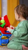 Sweet child playing with a bright lorry on the soft carpet. Toddler turns the car to take the toys out of the car basket. Blurred backdrop. Vertical video