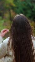arrière vue de une brunette fille en marchant par le parc dans l'automne. Dame se tourne autour pour une moment montrant une tasse dans sa main. flou toile de fond. verticale video