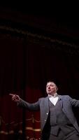 Actor performing on stage. Man in grey suit speaking with many gestures on red theatrical curtains background. View from below. Vertical video