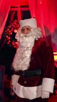 Bearded Santa Claus in cosy room with Christmas tree. Portrait of Santa looking on camera on the background of a New year tree and a fireplace. Red evening light. Vertical video