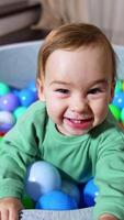 alegre contento niñito sonriente en general soportes en suave cuenca lleno con vistoso pelotas. alegre niño molesto a alcanzar el cámara. cerca arriba. vertical video