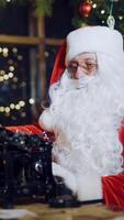 Santa Claus is sitting and writing letters on the typewriter for children on the background of Christmas tree with toys and garlands near window in the room. Preparation for New Year Vertical video