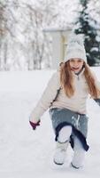 A little girl in warm clothes is descending from the hill and touching the snow. The child is scattering snow by her hands on the background of snowfall in the park. Slow motion Vertical video