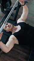 Strong healthy bodybuilder exercising in gym. Handsome guy lifting barbell while laying on a simulator. Top view. Sports concept. Vertical video
