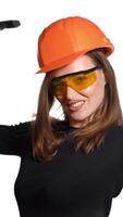 A pretty woman with orange helmet and eyeglasses is posing with a pickaxe in her hands on a white background in the studio. Profession of engineer Vertical video