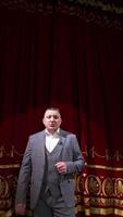 Man in gray costume on stage. Middle-aged actor talking on camera on luxury red curtains background in the theater. Slow motion. Vertical video
