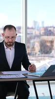 ocupado homem dentro terno trabalhando dentro escritório. meia idade empreendedor dentro óculos sentado às mesa com muitos papéis e uma computador portátil e trabalhando seriamente. vertical video