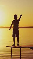A little boy is standing on the masonry near the bench and doing physical exercises. The child is raising hands up alternately on the background of the river at sunset. Vertical video